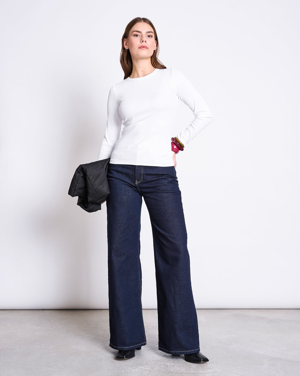 A woman stands against a white background, wearing the jan-n-june RIB LONGSLEEVE RAVA WHITE top, dark wide-leg jeans, and black shoes, holding a black jacket in her left hand.
