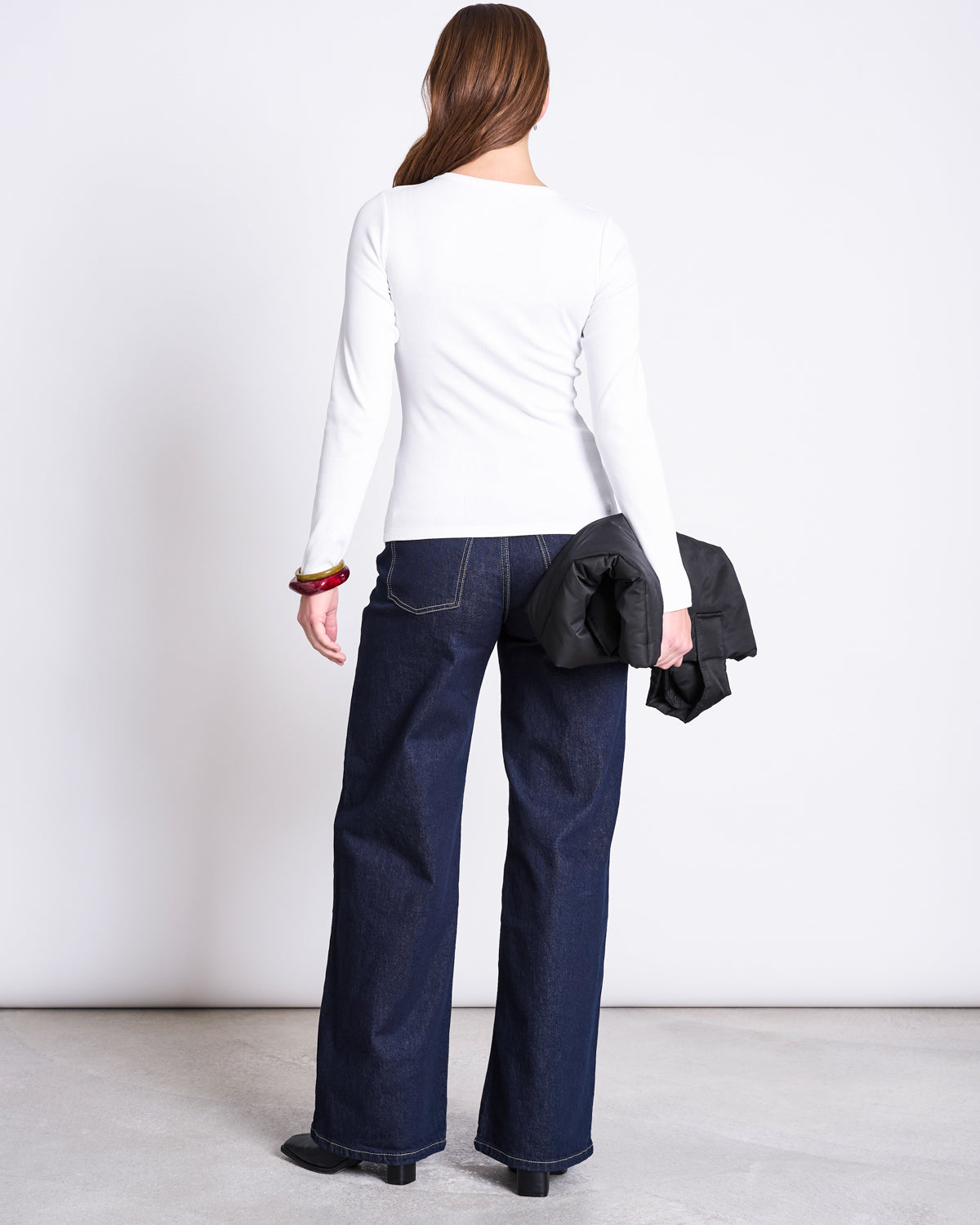 A woman with brown hair stands against a white background, wearing the jan-n-june RIB LONGSLEEVE RAVA WHITE top, dark wide-leg jeans, black shoes, and holding a black jacket in one hand.
