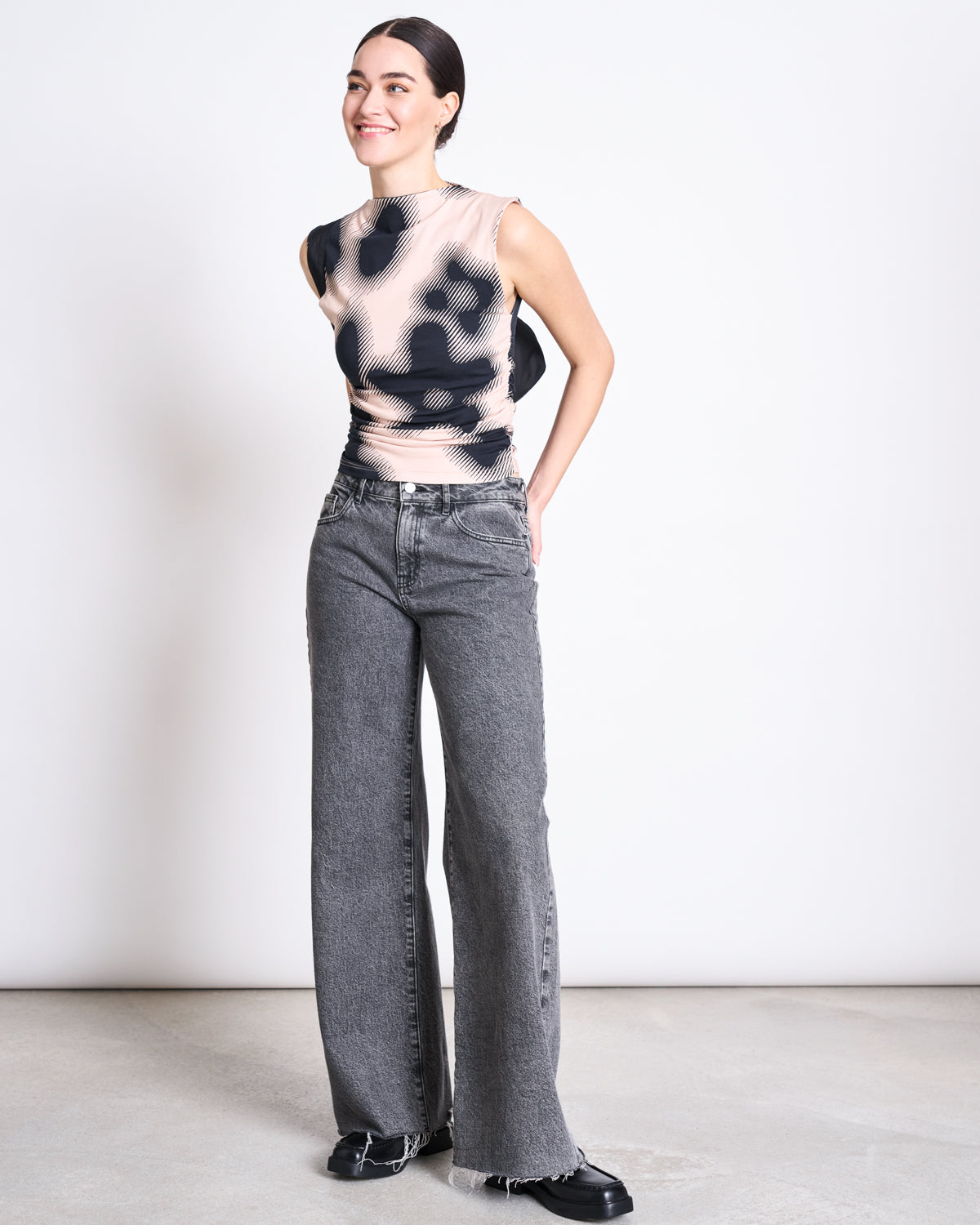 A woman smiles against a plain background, wearing the jan-n-june RUFFLE TOP RAFO DIGITAL FLOWER PRINT GOTS, paired with high-waisted gray wide-leg jeans and black shoes. Her dark hair is pulled back and her hands are behind her back.