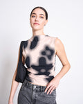 A woman with dark hair tied back wears the jan-n-june RUFFLE TOP RAFO DIGITAL FLOWER PRINT GOTS in organic cotton jersey, featuring a high neckline and abstract beige and black print, paired with high-waisted gray jeans against a white background.