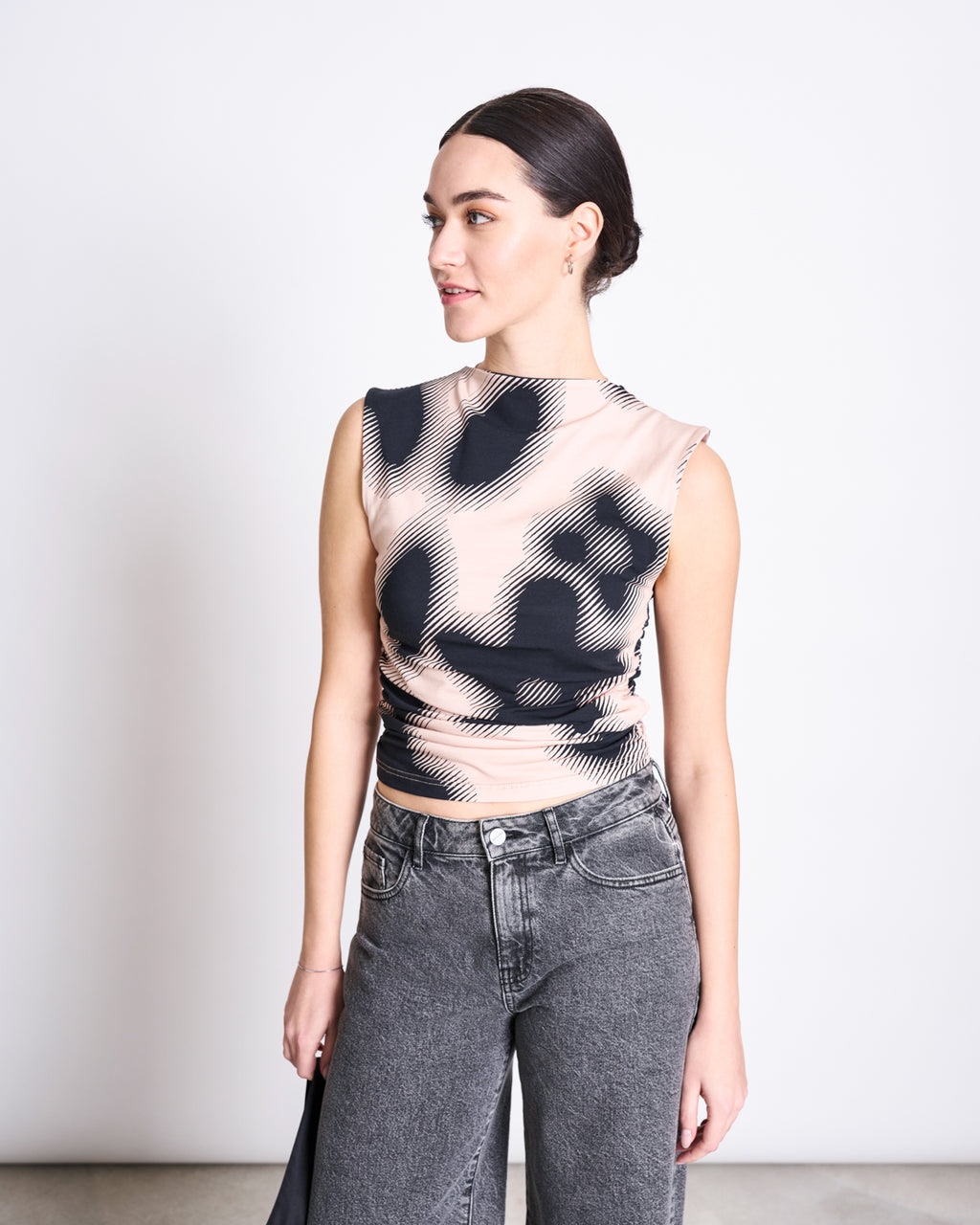 A woman with dark hair pulled back models the jan-n-june RUFFLE TOP RAFO DIGITAL FLOWER PRINT GOTS, a sleeveless high-neck ruffle top with abstract black and beige swirls, styled with high-waisted gray jeans against a white background.