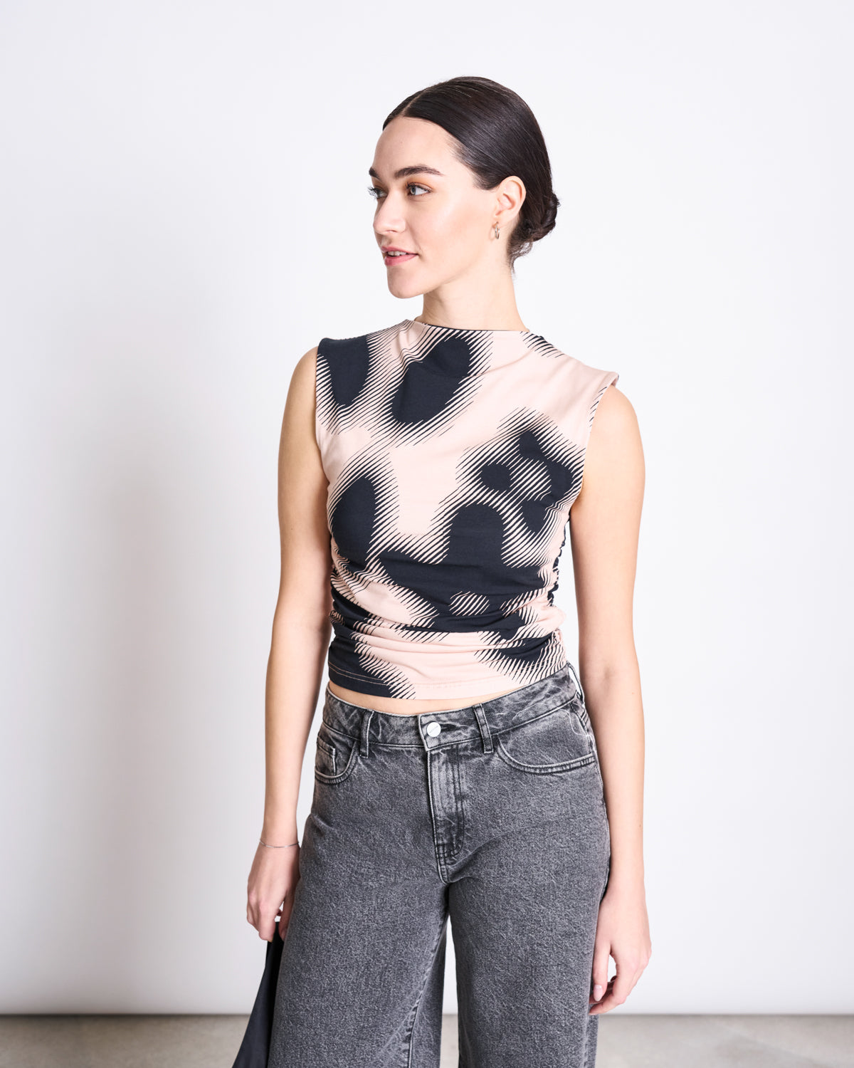 A woman with dark hair pulled back models the jan-n-june RUFFLE TOP RAFO DIGITAL FLOWER PRINT GOTS, a sleeveless high-neck ruffle top with abstract black and beige swirls, styled with high-waisted gray jeans against a white background.