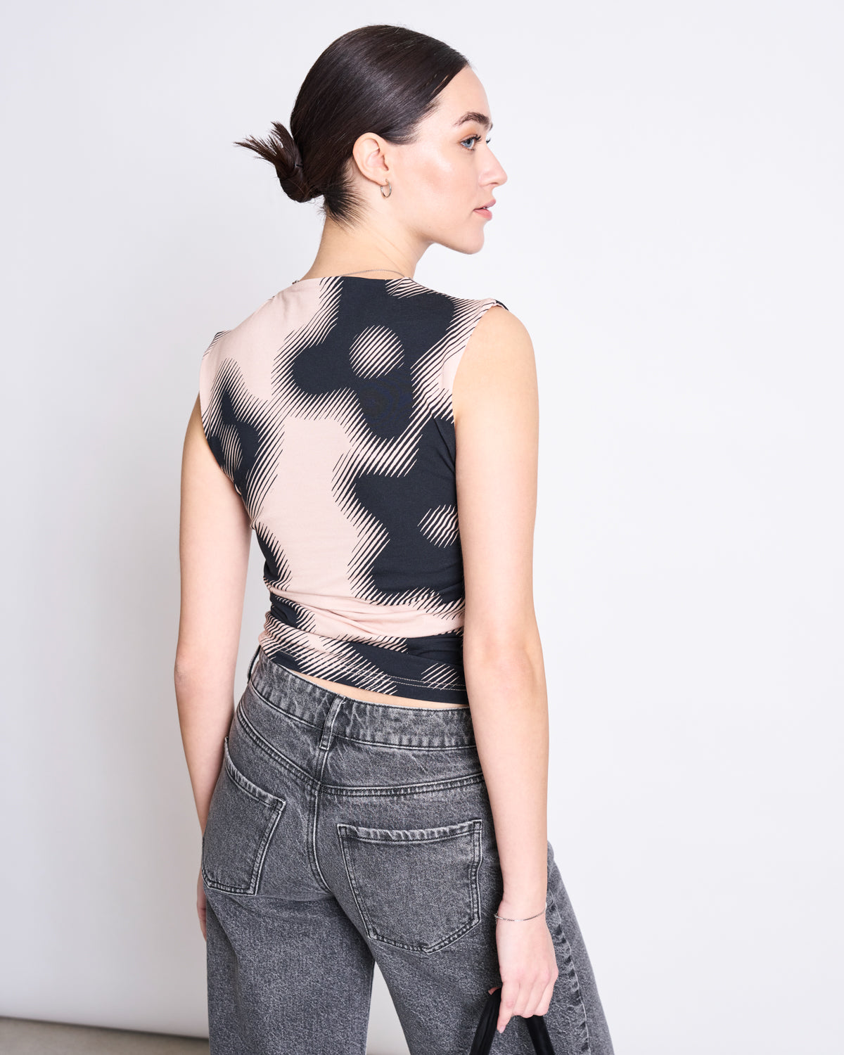 A woman with dark hair in a low bun wears the jan-n-june RUFFLE TOP RAFO DIGITAL FLOWER PRINT GOTS with high-waisted gray jeans, standing with her back to the camera against a plain, light background.