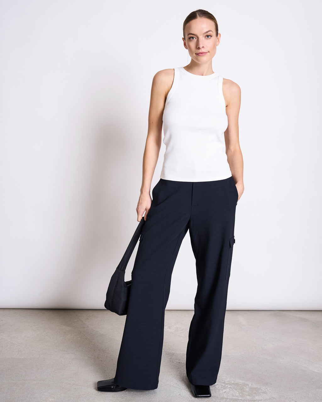 A woman poses against a plain background in the jan-n-june ORGANIC COTTON TANK TOP PORTO LONG WHITE GOTS and loose black pants. She carries a black shoulder bag, has one hand in her pocket, and her hair is pulled back—perfect for everyday wear.