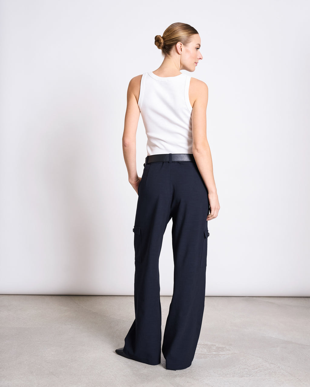 A woman with her hair in a neat bun wears the jan-n-june ORGANIC COTTON TANK TOP PORTO LONG WHITE GOTS, a slim fit ribbed tank, paired with dark wide-leg trousers. She stands on a light floor against a plain white background.
