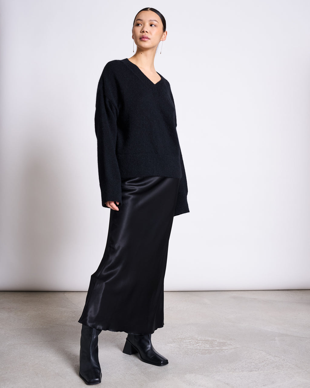 A person stands indoors against a plain light wall and concrete floor, wearing the jan-n-june REC. MOHAIR V-NECK JUMPER YICA BLACK, paired with a long black satin skirt and black heeled ankle boots.