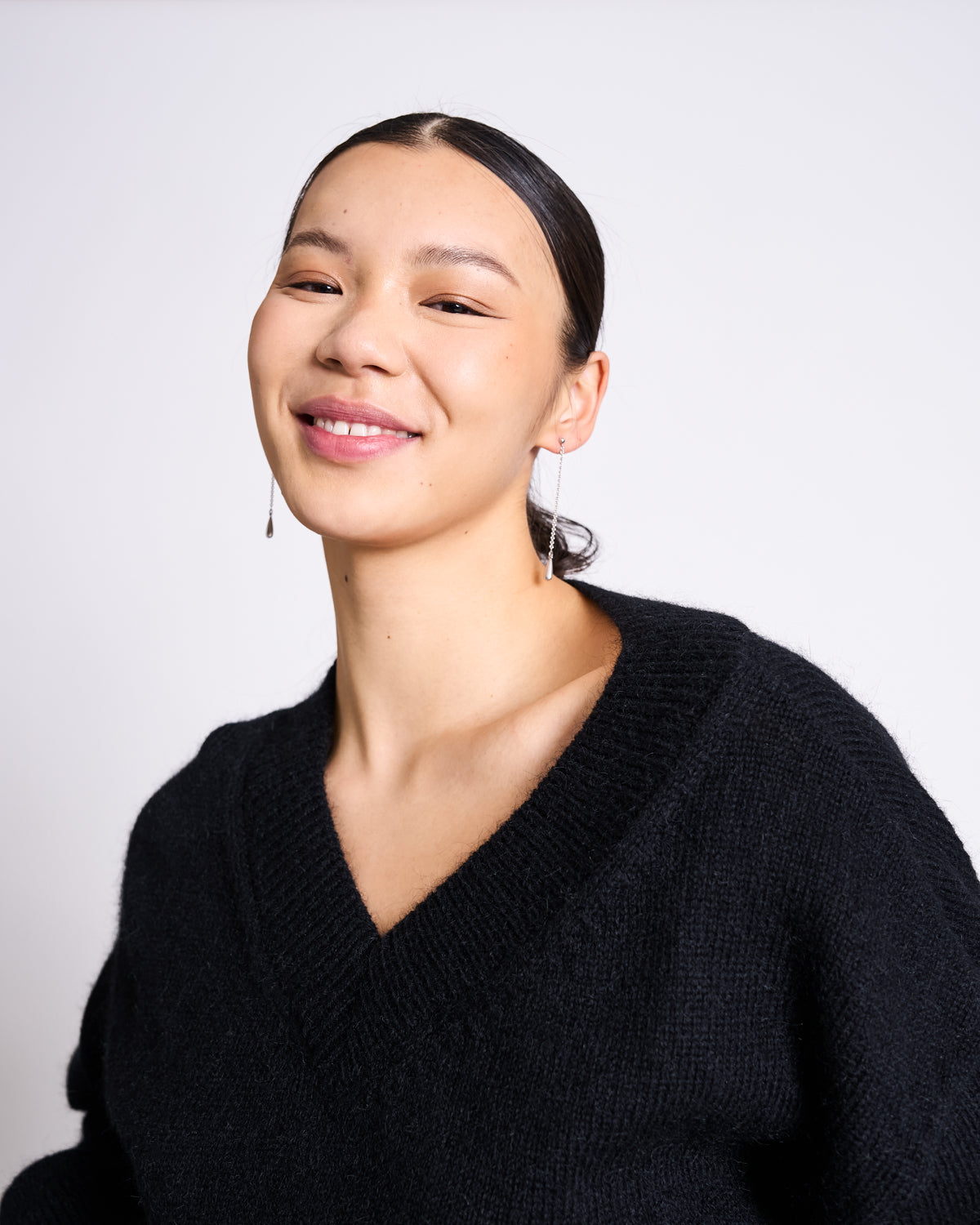 A young woman with straight dark hair pulled back, wearing the jan-n-june REC. MOHAIR V-NECK JUMPER YICA BLACK and long earrings, smiles softly while standing against a plain light background.