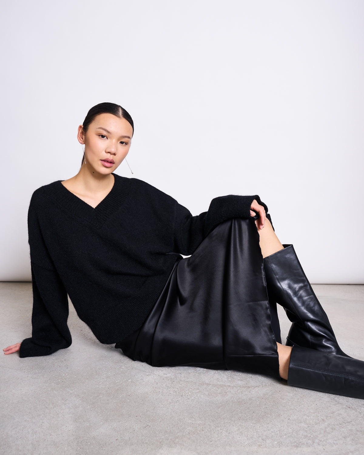A woman with straight dark hair sits on the floor by a white wall, wearing the jan-n-june REC. MOHAIR V-NECK JUMPER YICA BLACK, a black satin skirt, and knee-high black leather boots, looking relaxed with one knee raised.