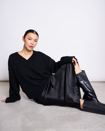 A woman with straight dark hair sits on the floor by a white wall, wearing the jan-n-june REC. MOHAIR V-NECK JUMPER YICA BLACK, a black satin skirt, and knee-high black leather boots, looking relaxed with one knee raised.