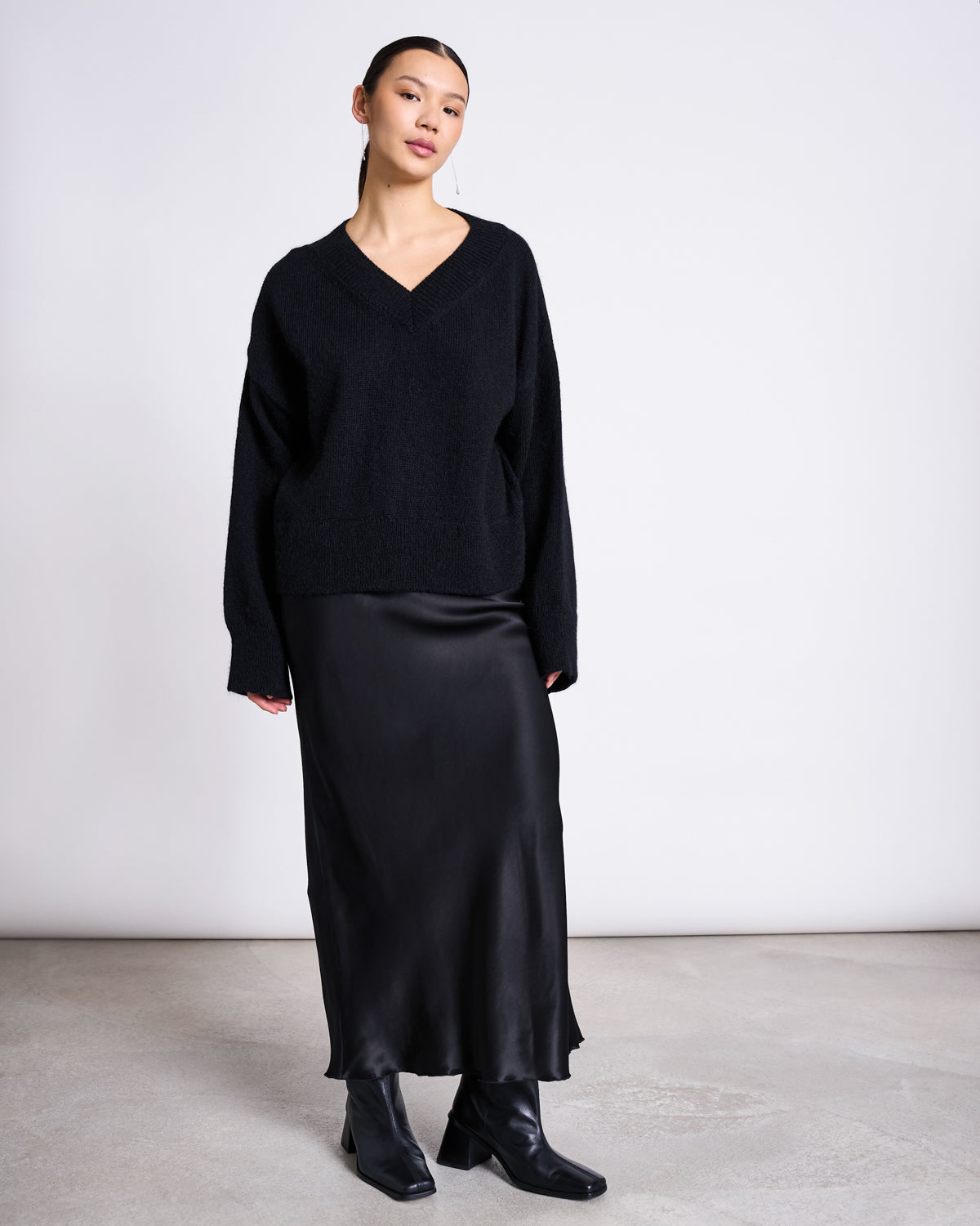 In a minimal studio, a woman wears the jan-n-june REC. MOHAIR V-NECK JUMPER YICA BLACK with a long satin skirt and ankle boots. Her hair is tied back as she gazes sideways, capturing an effortlessly stylish vibe.