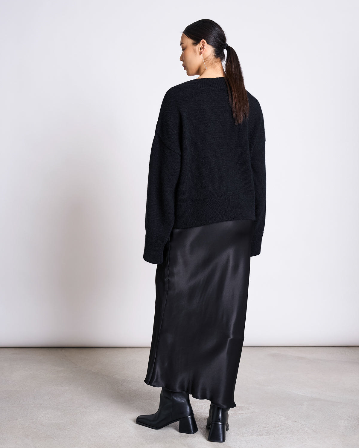 A woman with dark hair in a low ponytail stands facing away, wearing the jan-n-june REC. MOHAIR V-NECK JUMPER YICA BLACK, a long black satin skirt, and black ankle boots against a plain white wall on a light floor.