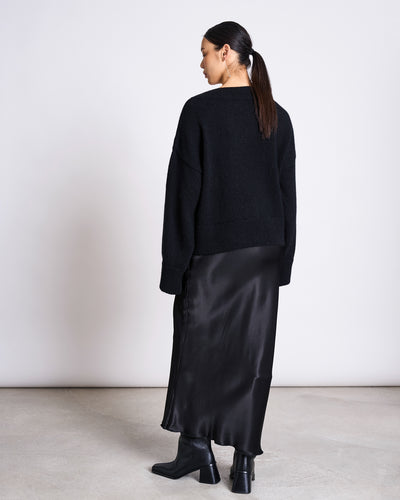 A woman with dark hair in a low ponytail stands facing away, wearing the jan-n-june REC. MOHAIR V-NECK JUMPER YICA BLACK, a long black satin skirt, and black ankle boots against a plain white wall on a light floor.