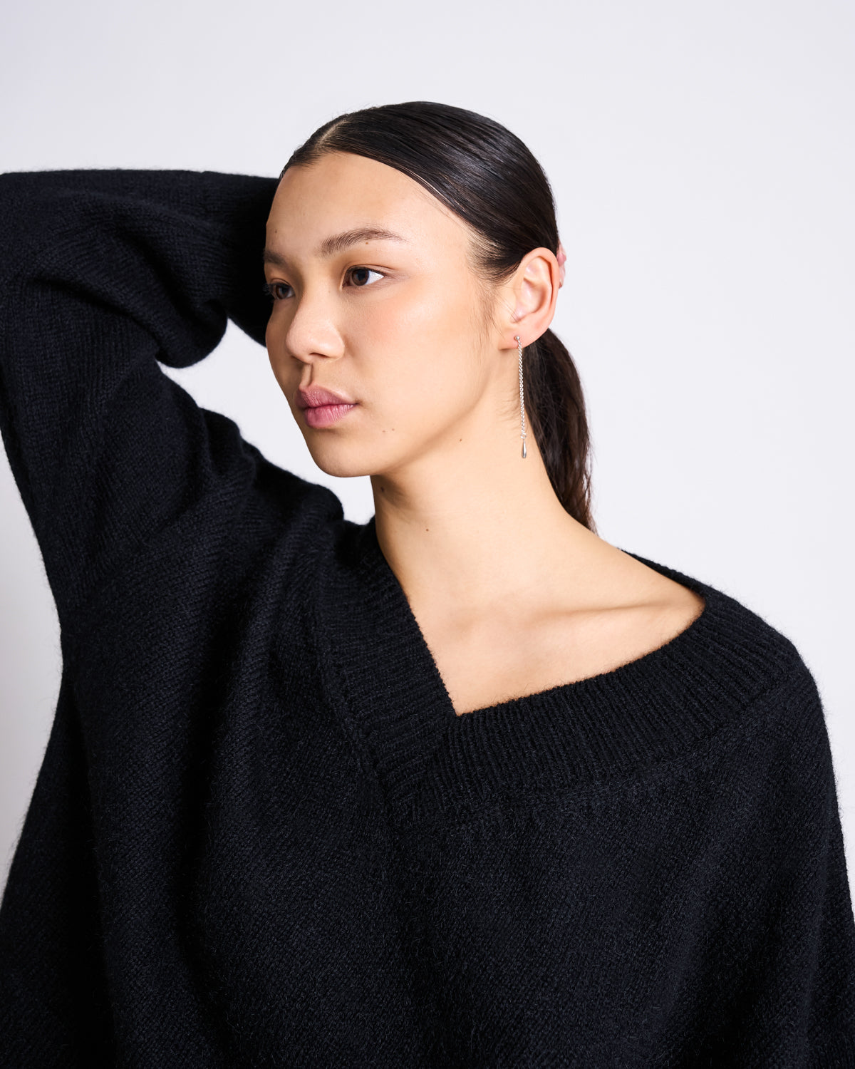A woman with straight dark hair pulled back poses with one arm behind her head, wearing the jan-n-june REC. MOHAIR V-NECK JUMPER YICA BLACK and a single dangling earring against a plain white background.
