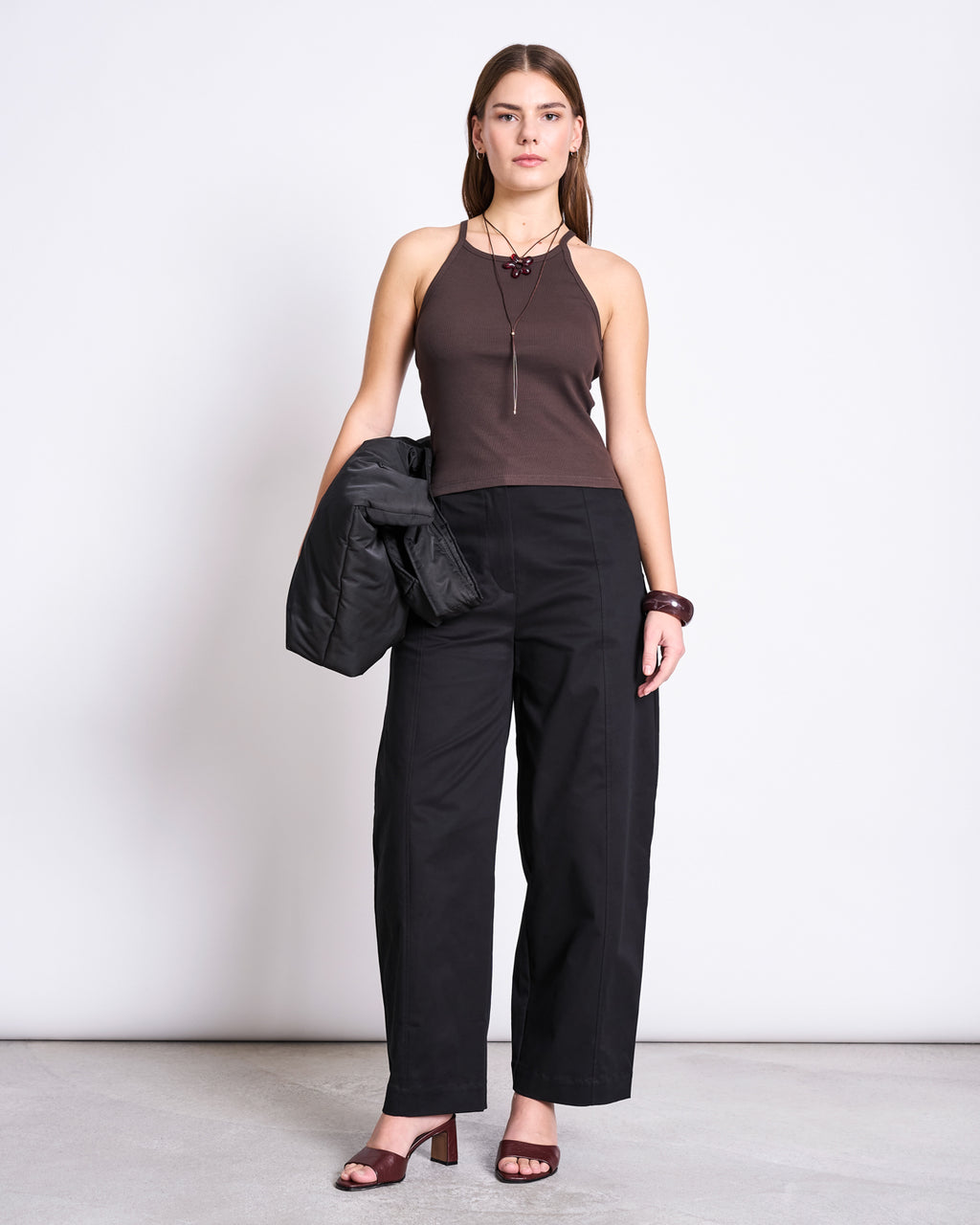 A woman with a neutral expression stands against a plain background wearing the jan-n-june RIB TOP STEM MUD, black wide-leg pants, brown heeled sandals, jewelry, and holds a black jacket in one hand.