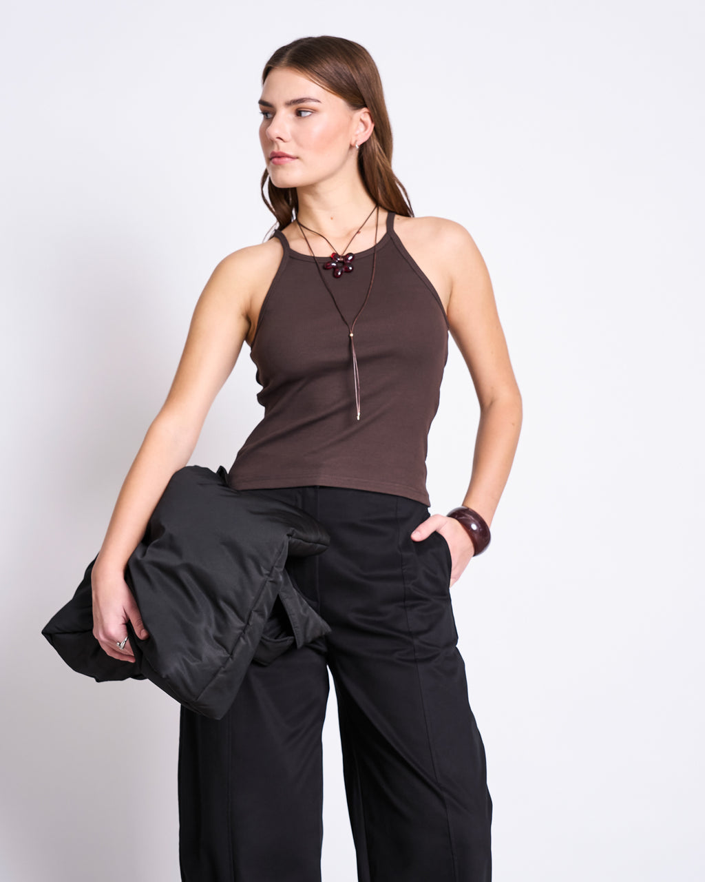 A woman stands against a plain background wearing the jan-n-june RIB TOP STEM MUD, black wide-leg pants, and a necklace. She holds a black jacket in one hand and gazes to the side with a neutral expression.