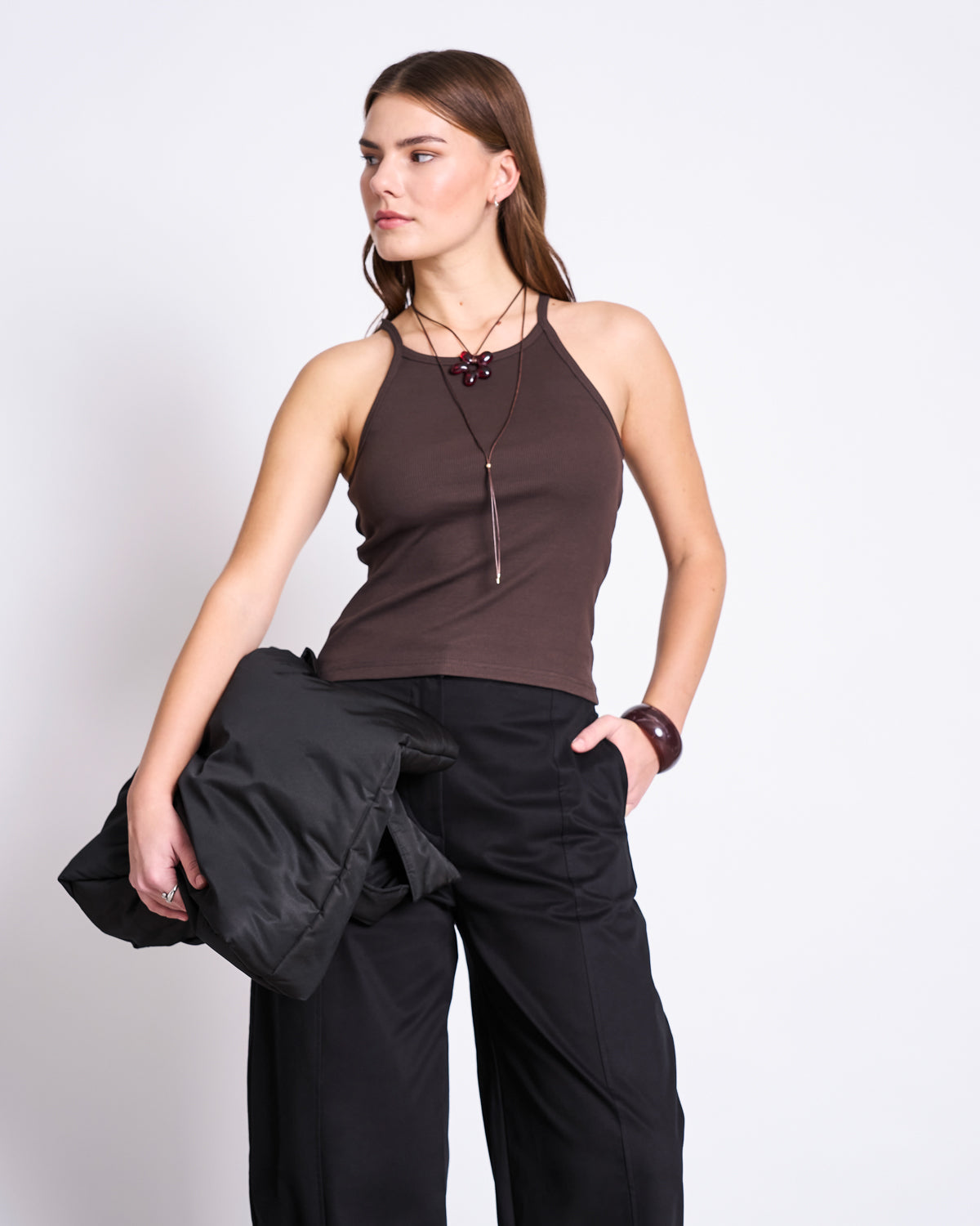 A woman stands against a plain background wearing the jan-n-june RIB TOP STEM MUD, black wide-leg pants, and a necklace. She holds a black jacket in one hand and gazes to the side with a neutral expression.