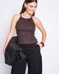 A woman with straight brown hair smiles while wearing the jan-n-june RIB TOP STEM MUD, an organic cotton rib top in dark brown, paired with black pants. She holds a black jacket and wears a necklace and bracelet against a plain white background.