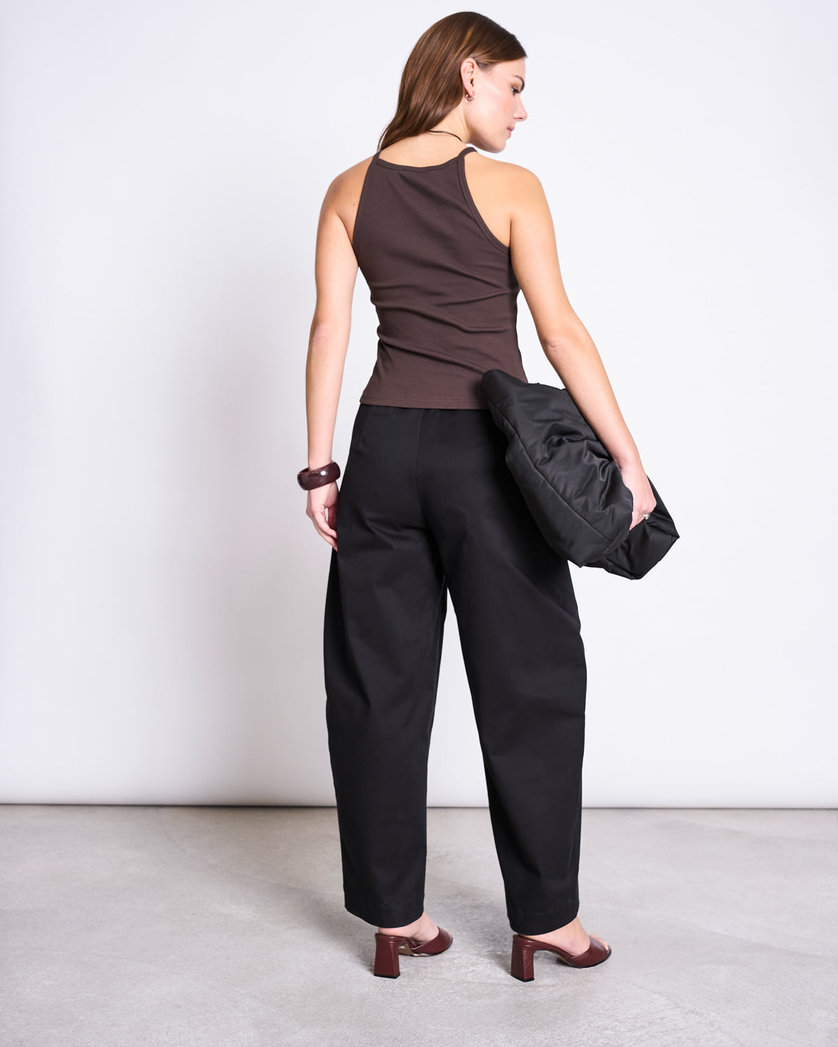 A woman faces away in jan-n-june’s RIB TOP STEM MUD, paired with black wide-leg pants and brown heeled sandals. She holds a black bag under her arm and wears a chunky bracelet on her right wrist against a plain, light background.