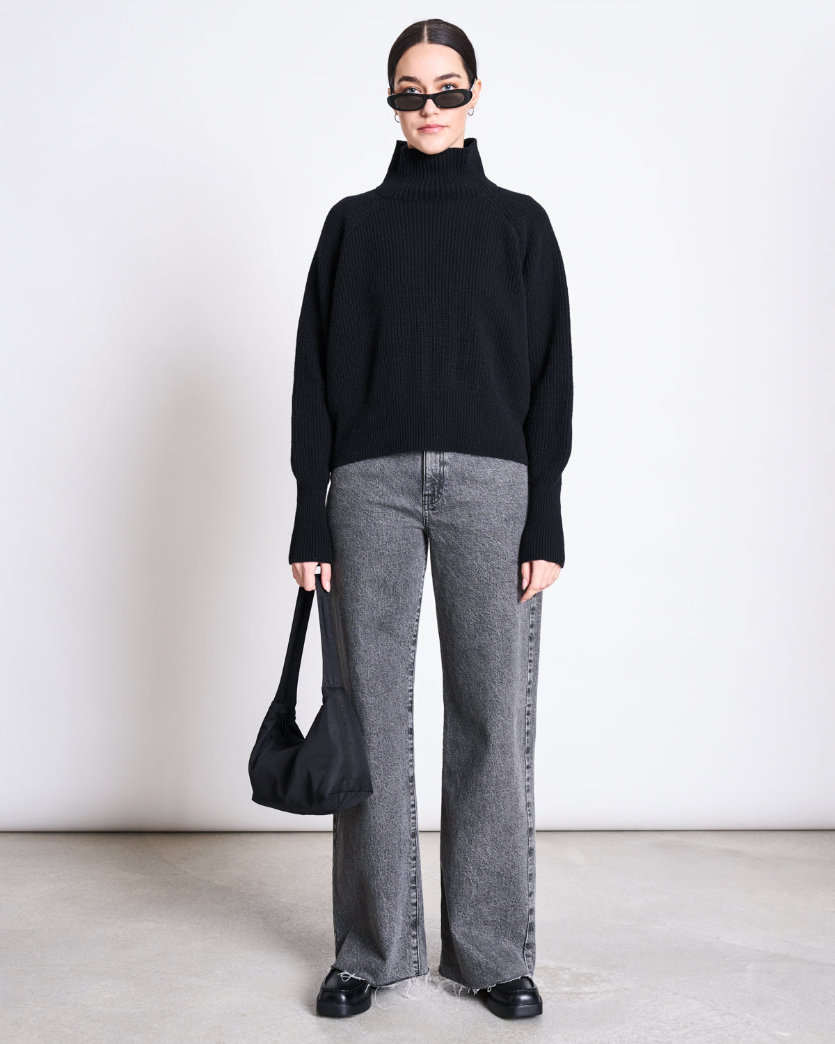 A person stands against a white background wearing jan-n-junes HIGH-NECK JUMPER WOOLY OLA BLACK GOTS, paired with gray wide-leg jeans, black shoes and sunglasses, and holding a black handbag.