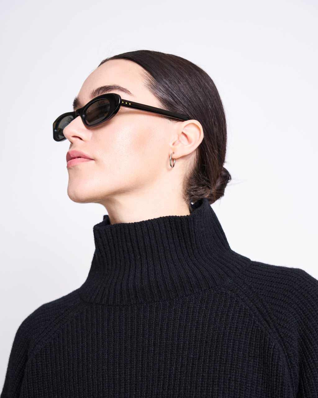 Woman with dark hair in a low bun wears black cat-eye sunglasses, small hoop earrings, and the jan-n-june HIGH-NECK JUMPER WOOLY OLA BLACK GOTS, posing against a plain white background.