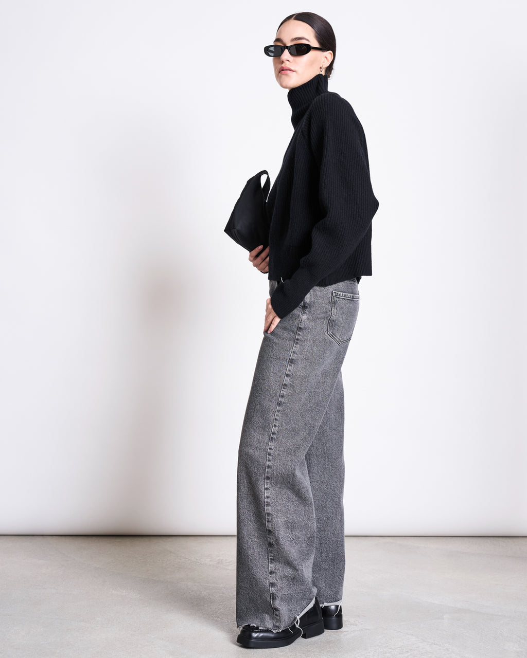 A woman wearing the jan-n-june HIGH-NECK JUMPER WOOLY OLA BLACK GOTS, black sunglasses, gray wide-leg jeans, black shoes, and holding a small black handbag stands sideways against a plain white background.