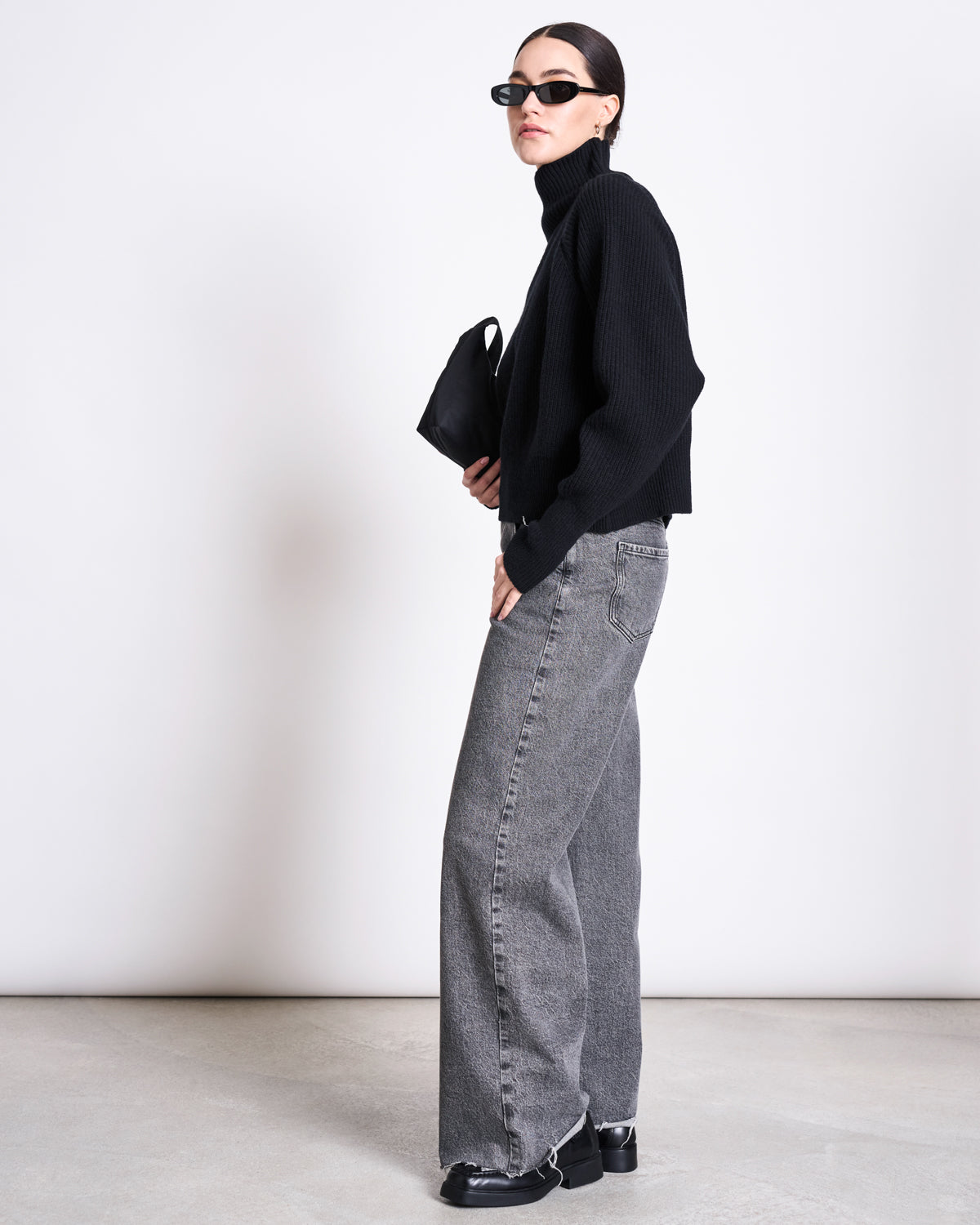 A woman wearing the jan-n-june HIGH-NECK JUMPER WOOLY OLA BLACK GOTS, black sunglasses, gray wide-leg jeans, black shoes, and holding a small black handbag stands sideways against a plain white background.
