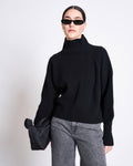 A woman in the jan-n-june HIGH-NECK JUMPER WOOLY OLA BLACK GOTS, black sunglasses, gray jeans, and holding a black clutch stands against a plain white background.