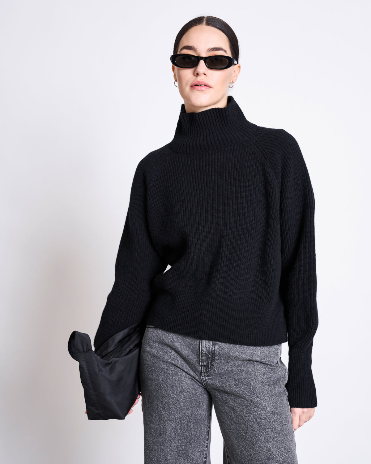 A woman in the jan-n-june HIGH-NECK JUMPER WOOLY OLA BLACK GOTS, black sunglasses, gray jeans, and holding a black clutch stands against a plain white background.