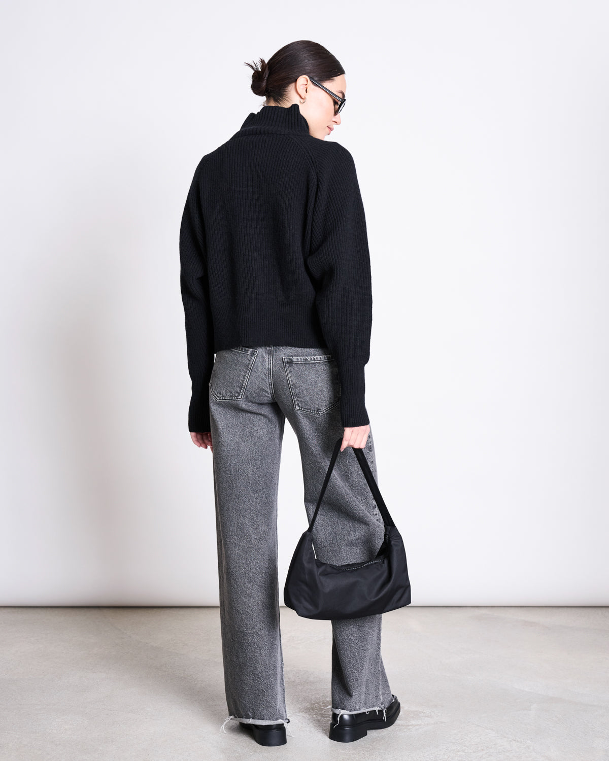 A person stands with their back to the camera in a plain white setting, wearing the jan-n-june HIGH-NECK JUMPER WOOLY OLA BLACK GOTS, gray high-waisted jeans, black shoes, sunglasses, and holding a black handbag by their side.