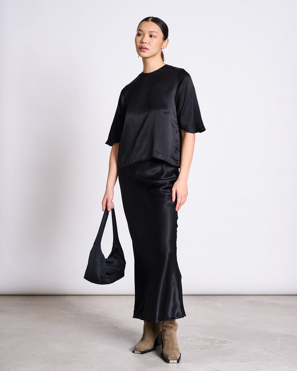 A woman stands against a plain light background, wearing the jan-n-june SILKY SHIRT NAMORA BLACK, a matching long black satin skirt made from ENKA Viscose, taupe ankle boots, and holding a black bag.