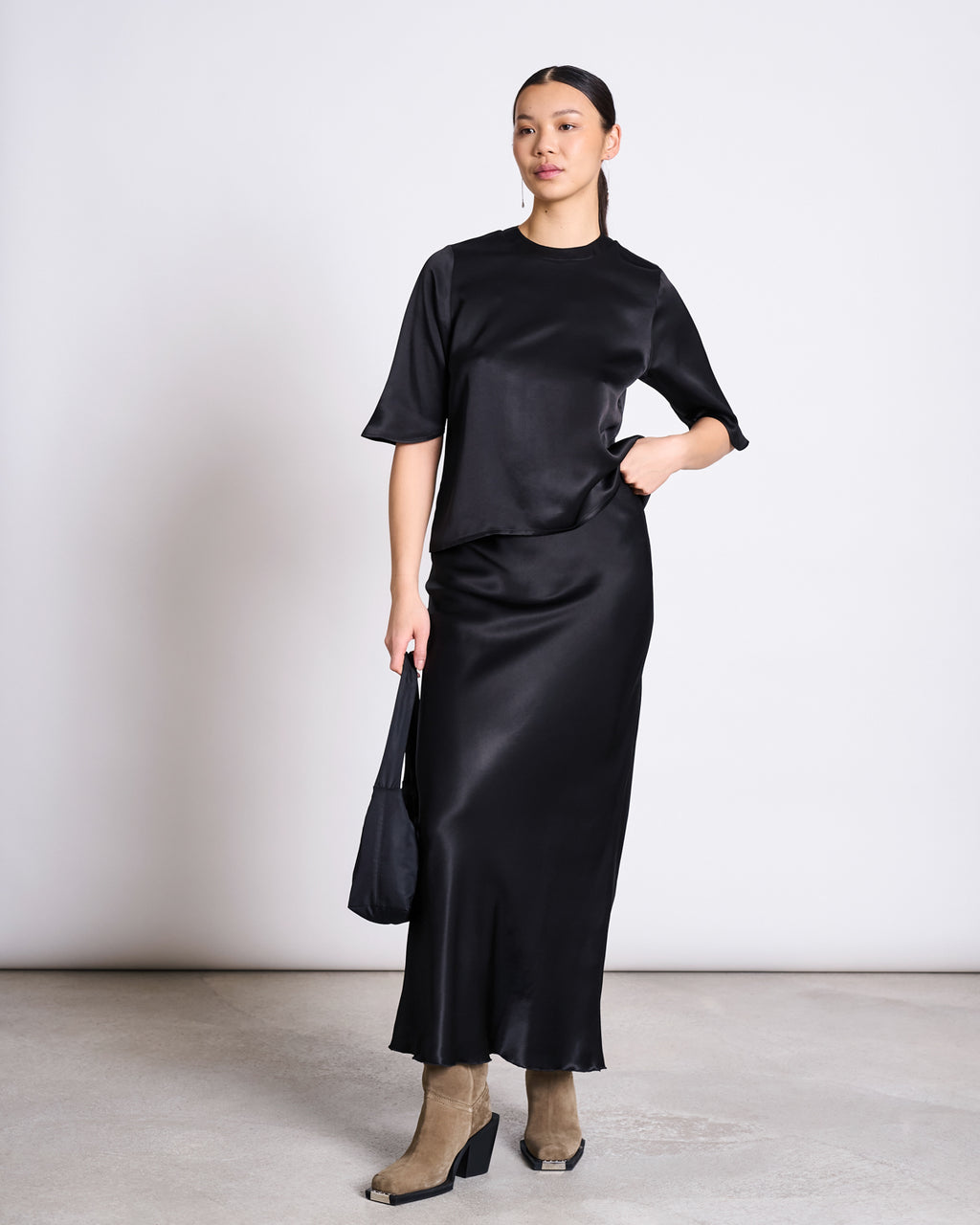 A woman stands against a plain background wearing the jan-n-june SILKY SHIRT NAMORA BLACK—made with ENKA Viscose—and a matching long skirt. She holds a small black bag, wears beige suede ankle boots, and has her hair tied back.