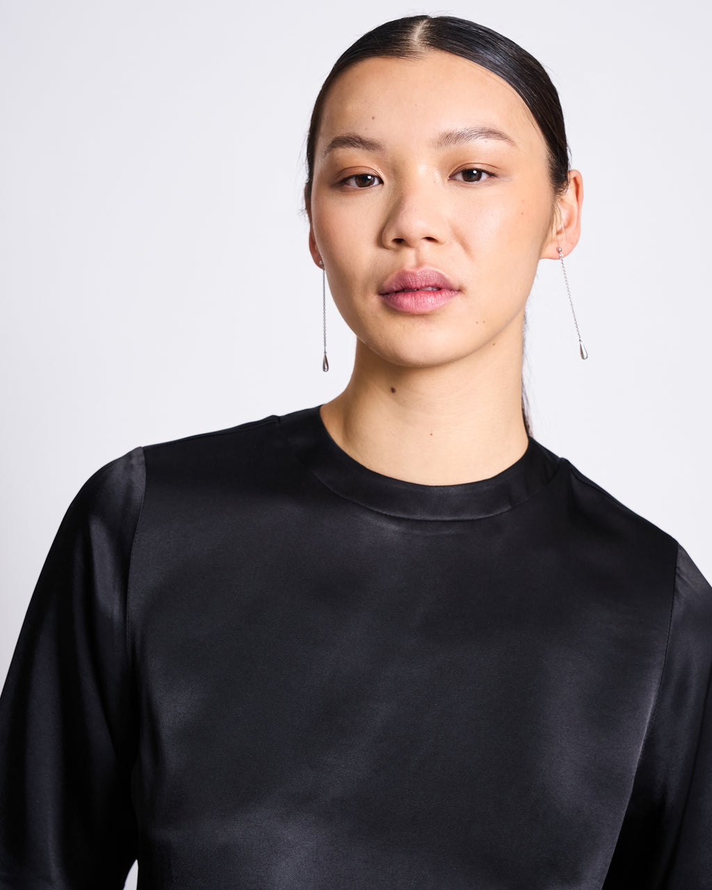 A woman with straight dark hair pulled back wears the jan-n-june SILKY SHIRT NAMORA BLACK and long, delicate drop earrings, posing against a plain white background.