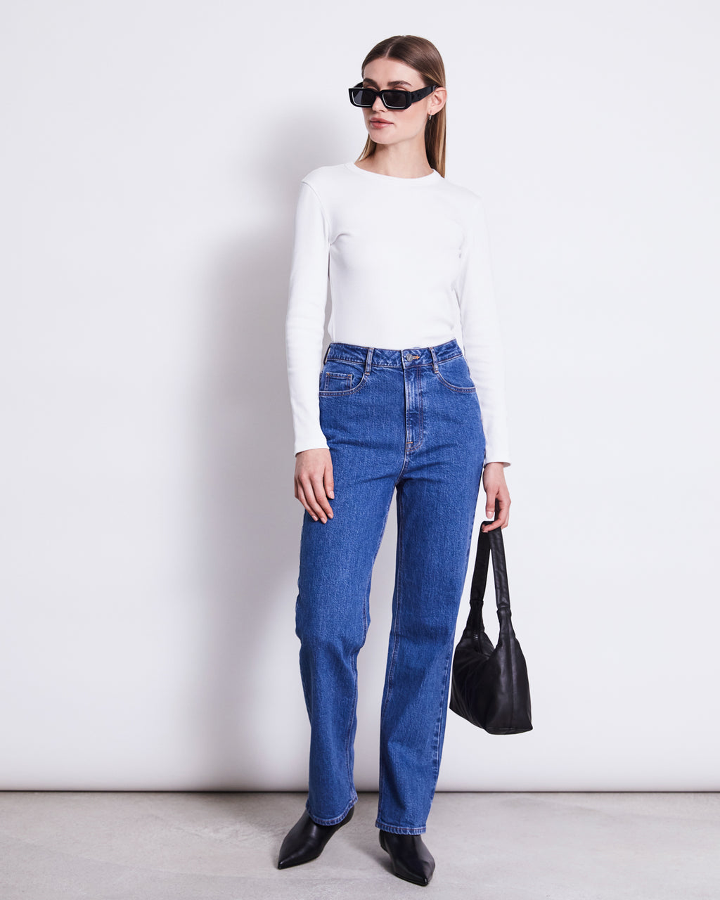 A woman in jan-n-junes RIB LONGSLEEVE MURI WHITE GOTS, blue high-waisted jeans, black sunglasses, pointed black shoes, and holding a black handbag stands against a plain white background.