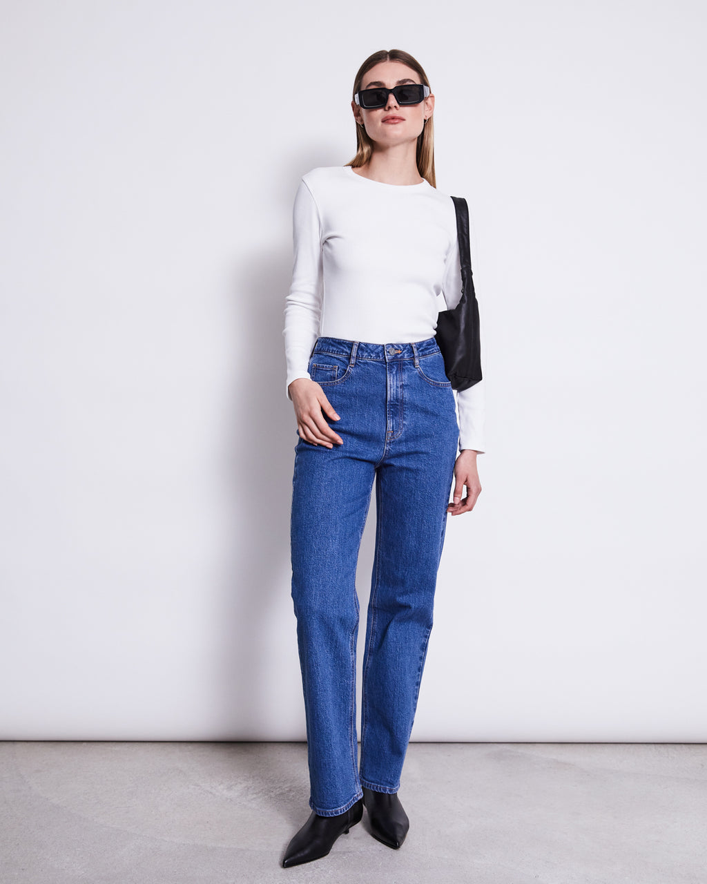 A woman stands against a plain white wall, wearing the jan-n-june RIB LONGSLEEVE MURI WHITE GOTS, blue high-waisted jeans, black pointed-toe shoes, sunglasses, and a black shoulder bag.