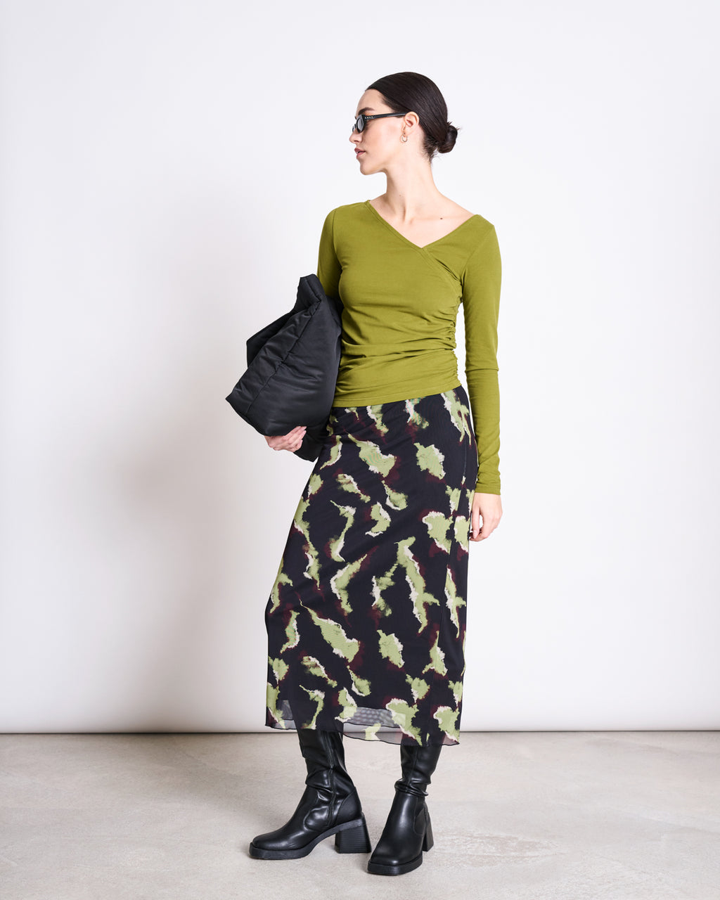 A woman in sunglasses and the jan-n-june ASYMMETRICAL LONGSLEEVE CASMA WOODY OLIVE GOTS top stands against a white wall, paired with a patterned midi skirt, black knee-high boots, and holding a black bag.