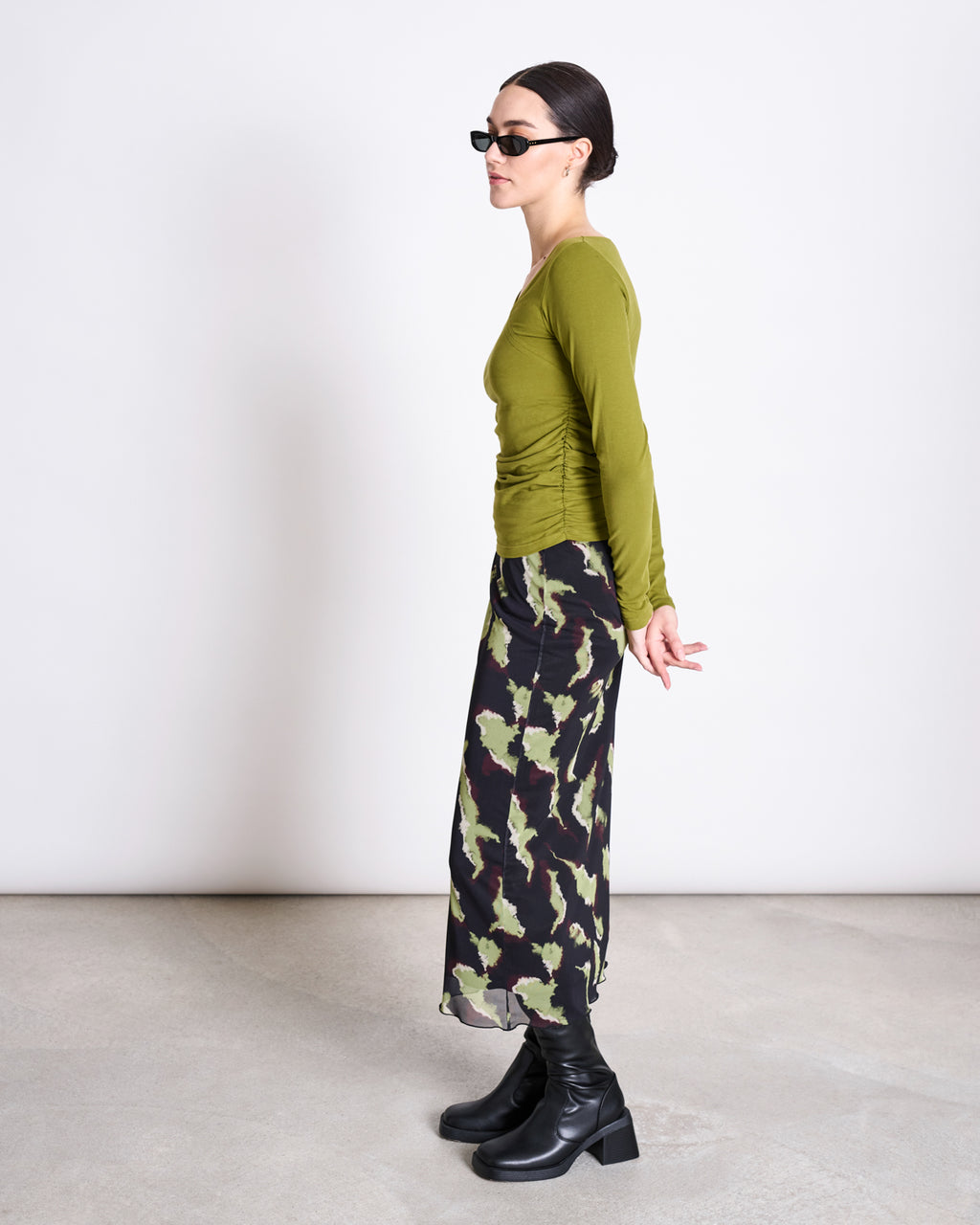 A woman stands in profile against a plain background, wearing jan-n-june’s ASYMMETRICAL LONGSLEEVE CASMA WOODY OLIVE GOTS top, sunglasses, a camouflage-patterned skirt, and black heeled boots with her hands behind her back.