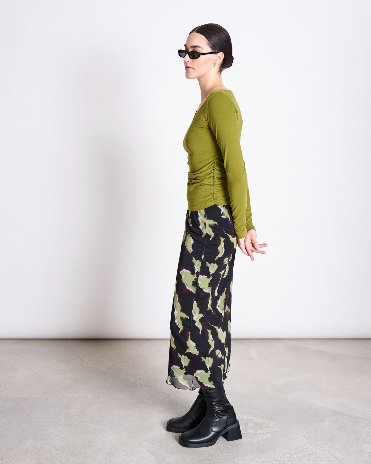 A woman stands in profile against a plain background, wearing jan-n-june’s ASYMMETRICAL LONGSLEEVE CASMA WOODY OLIVE GOTS top, sunglasses, a camouflage-patterned skirt, and black heeled boots with her hands behind her back.