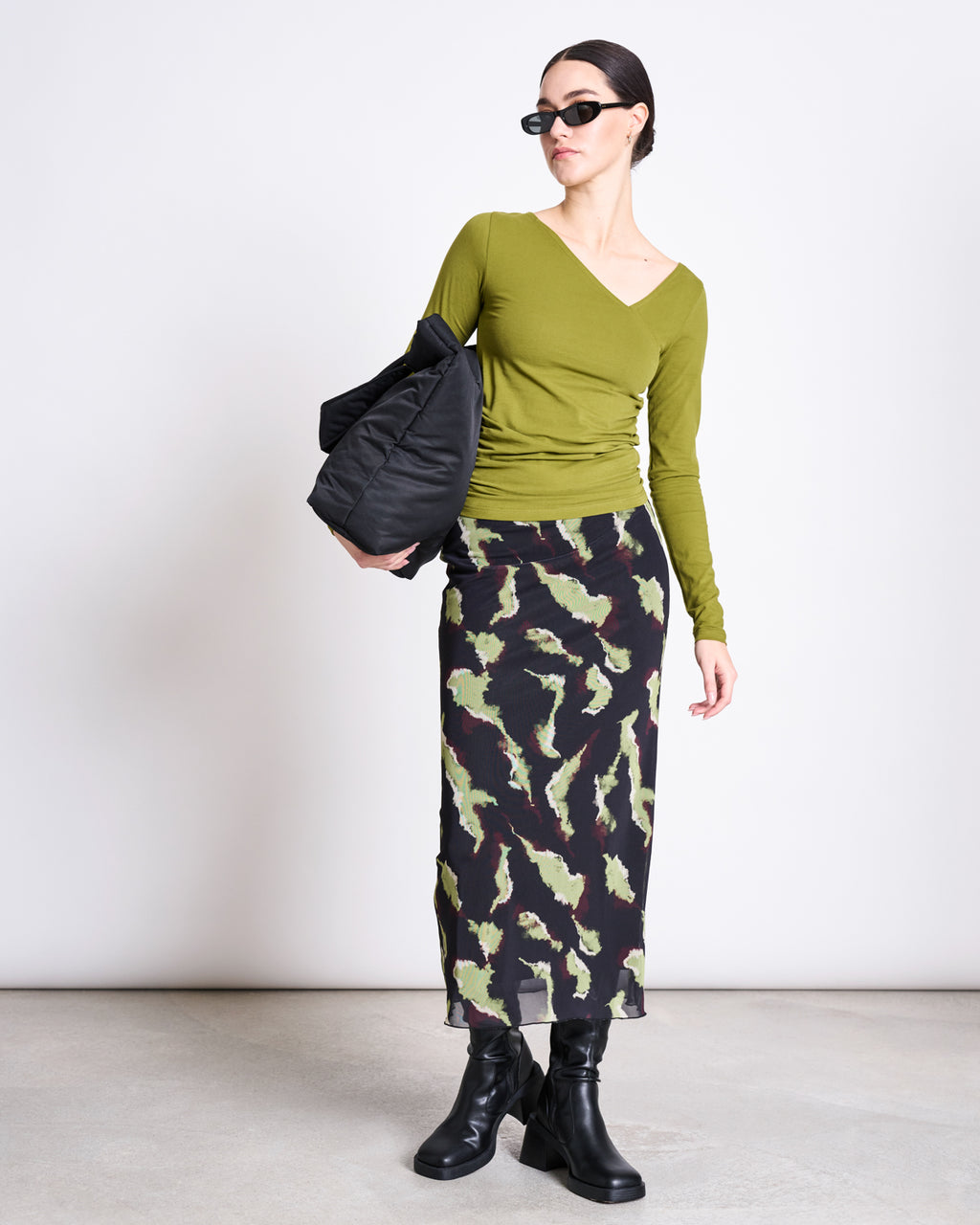 A woman stands against a plain light background wearing jan-n-junes ASYMMETRICAL LONGSLEEVE CASMA WOODY OLIVE GOTS, sunglasses, a black and green patterned skirt, black boots, and carrying a black bag.
