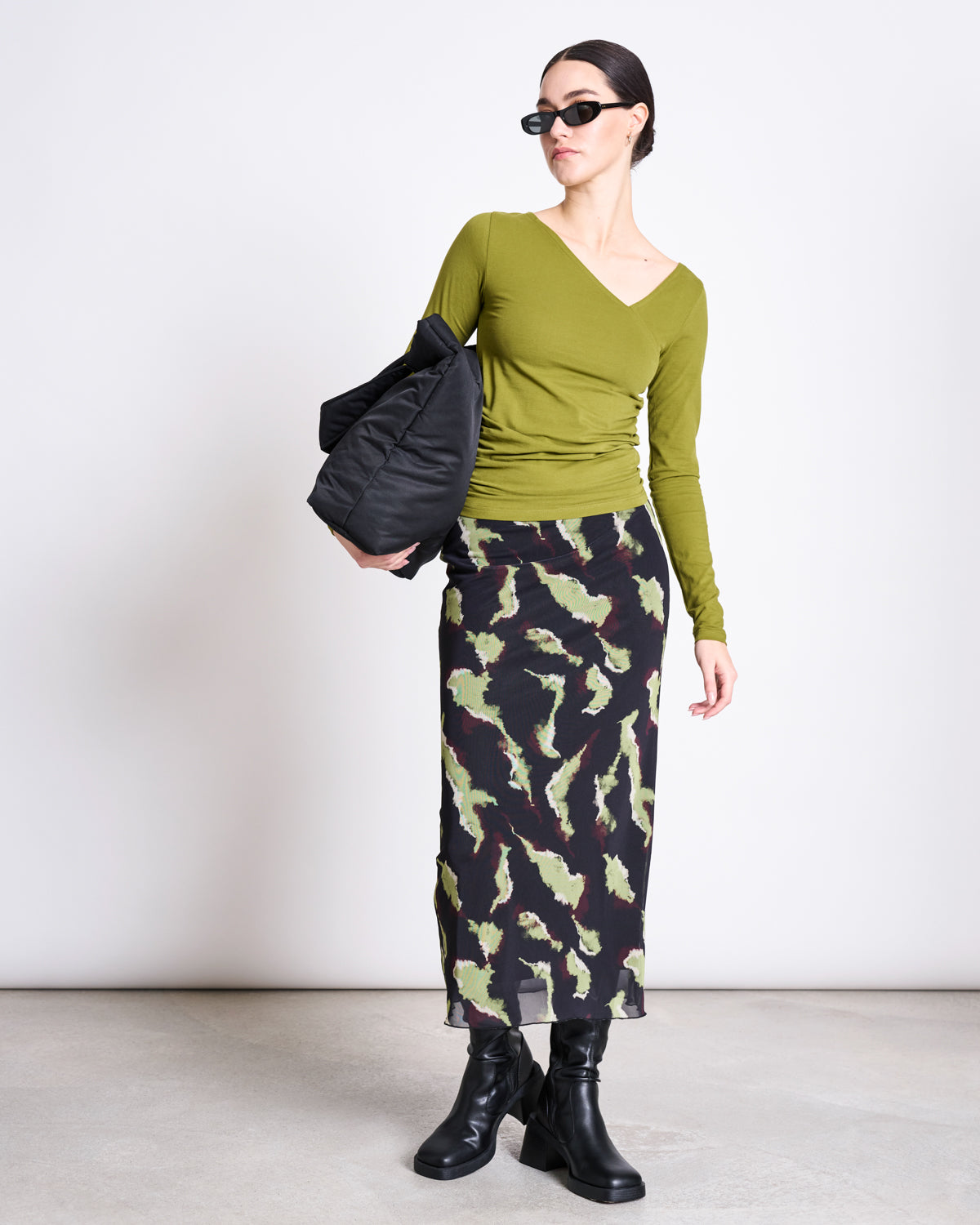 A woman wearing sunglasses, a fitted olive green top, black heeled boots, and holding a black quilted bag poses against a light background in the jan-n-june MESH MAXI SKIRT SMUK MOSSFIRE with an abstract print.