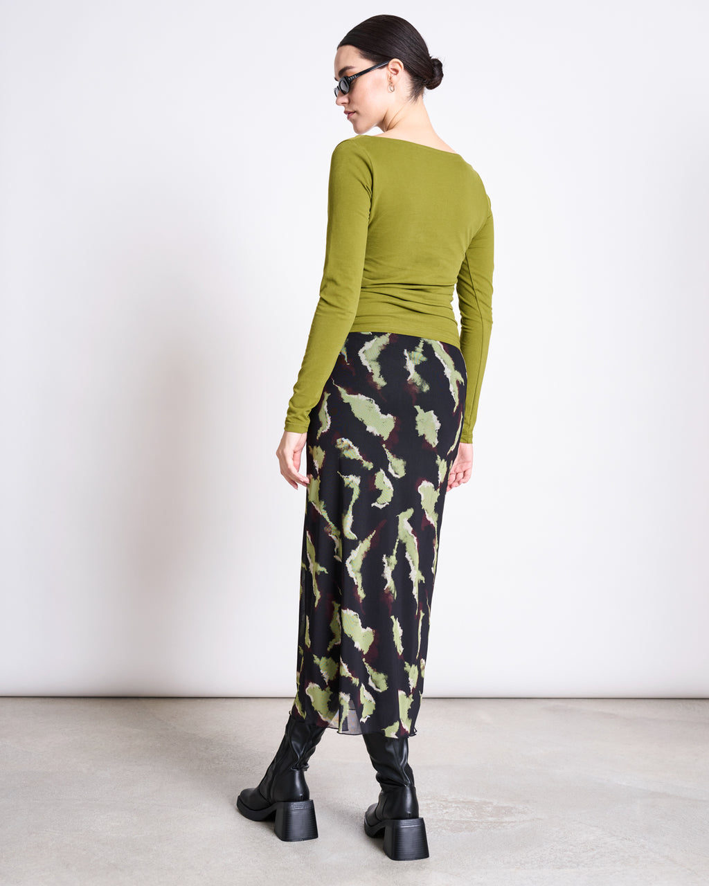 A woman faces away wearing the jan-n-june ASYMMETRICAL LONGSLEEVE CASMA WOODY OLIVE GOTS in organic cotton, paired with a black and green abstract skirt, chunky black boots, sunglasses, and her dark hair tied back.