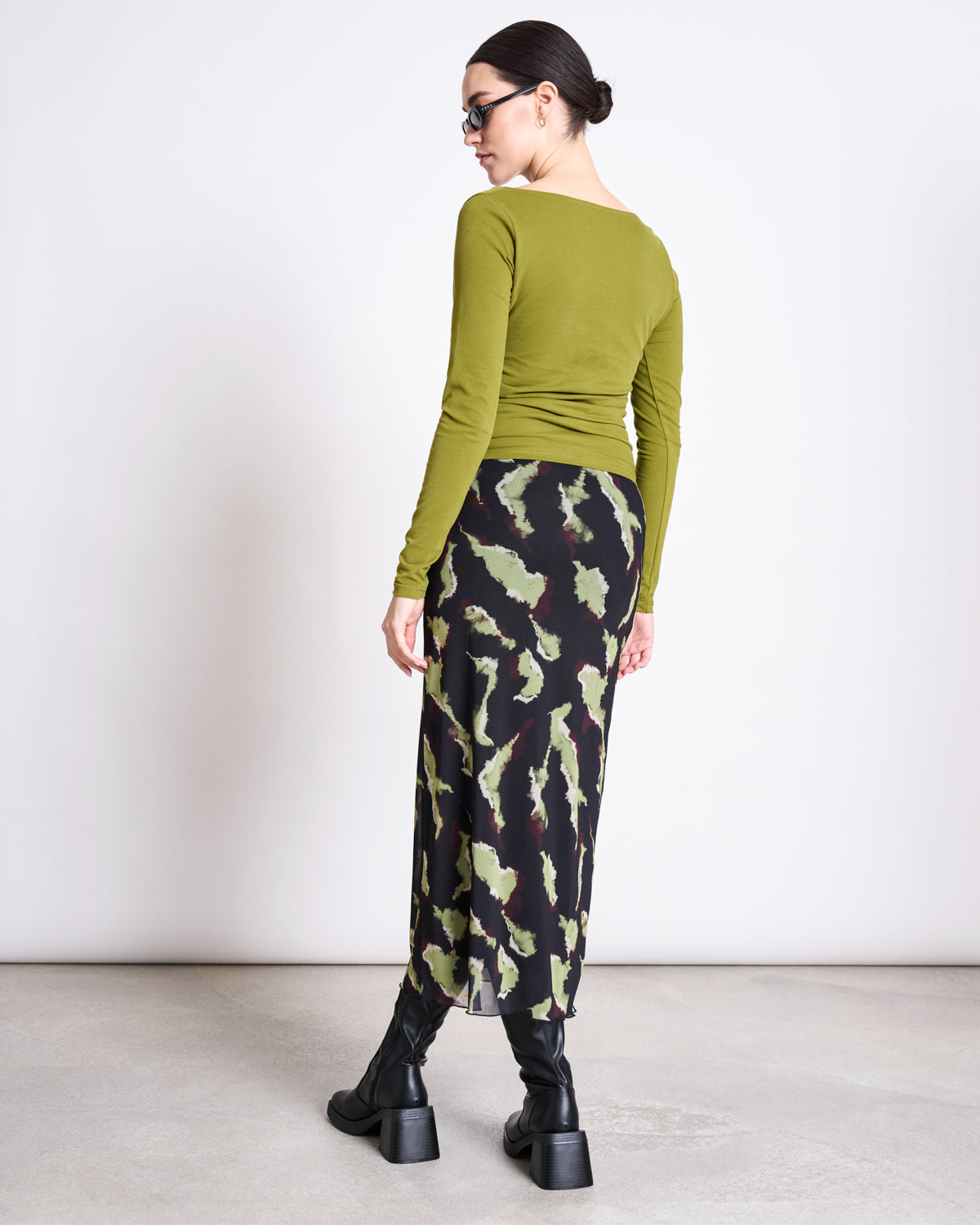 A woman faces away wearing the jan-n-june ASYMMETRICAL LONGSLEEVE CASMA WOODY OLIVE GOTS in organic cotton, paired with a black and green abstract skirt, chunky black boots, sunglasses, and her dark hair tied back.