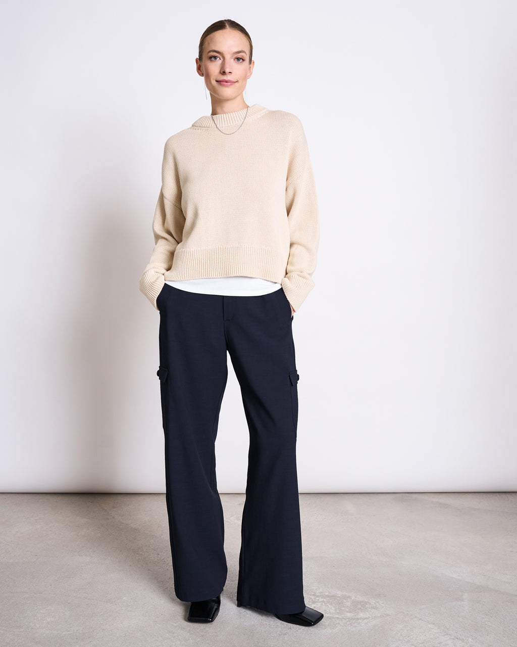 A person stands against a plain background, wearing the jan-n-june JUMPER YAGMUR BEIGE GOTS over a white shirt, paired with dark wide-leg pants with side pockets and black shoes, hands in pockets and a slight smile.