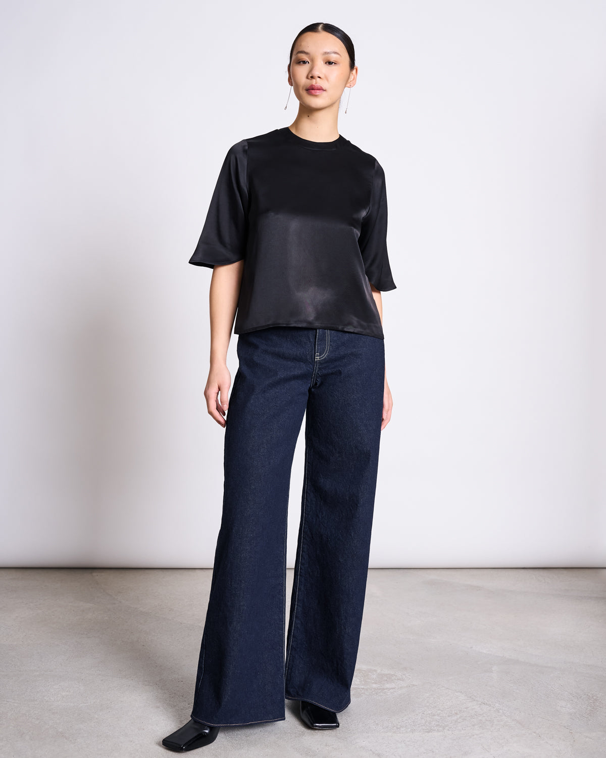 A woman stands in a minimalist room wearing a shiny black short-sleeve blouse, black square-toe shoes, and jan-n-junes WIDE LEG LOWRISE JEANS TACNA DARK BLUE DENIM GOTS. Her hair is pulled back, and she wears a neutral expression.