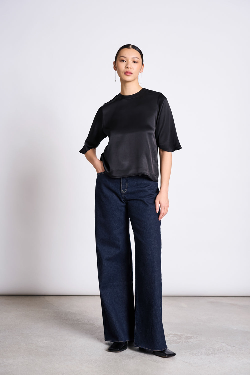 A person poses with one hand in their pocket, wearing jan-n-june’s WIDE LEG LOWRISE JEANS TACNA DARK BLUE DENIM GOTS and a loose black short-sleeve top, paired with black shoes; their straight hair is pulled back.