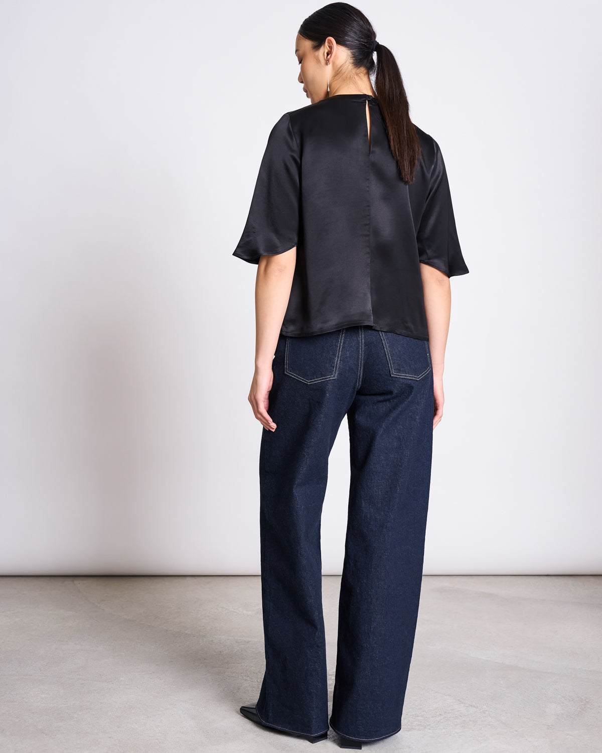 A woman with a low ponytail faces away from the camera, wearing the jan-n-june SILKY SHIRT NAMORA BLACK made from ENKA Viscose, paired with dark blue wide-leg jeans. The background is plain and light.