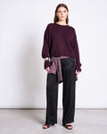 A woman stands against a plain background wearing the jan-n-june LIGHT CARDIGAN ASAS CHERRY LACQUER GOTS layered over a loose maroon sweater, black wide-leg pants, open-toe maroon shoes, and a striped scarf tied at her waist.