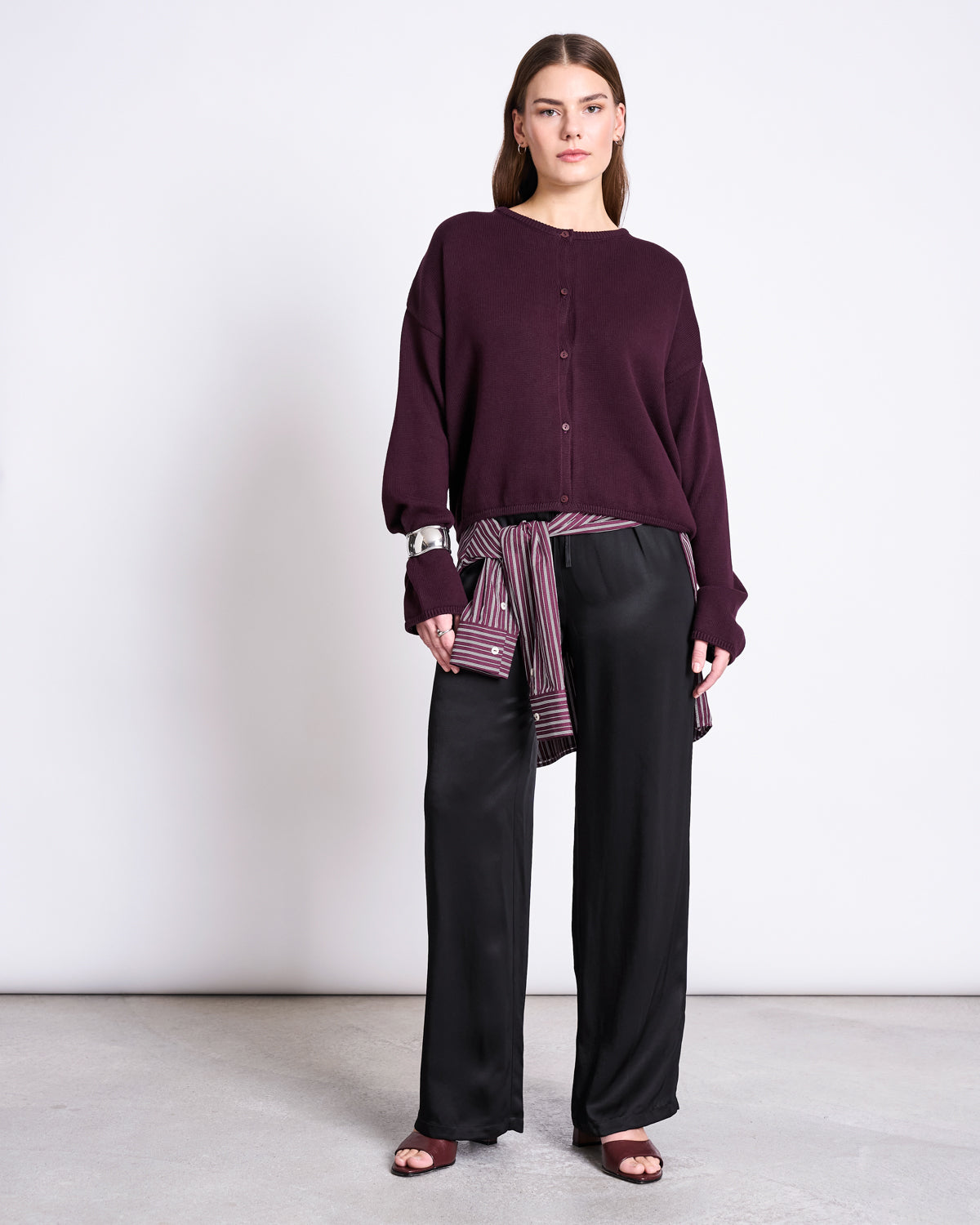A woman stands against a plain background wearing the jan-n-june LIGHT CARDIGAN ASAS CHERRY LACQUER GOTS layered over a loose maroon sweater, black wide-leg pants, open-toe maroon shoes, and a striped scarf tied at her waist.