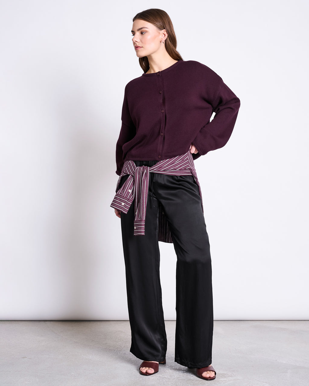 A woman stands against a plain background wearing jan-n-june's LIGHT CARDIGAN ASAS CHERRY LACQUER GOTS, black wide-leg pants, and a striped shirt tied around her waist. She looks to the side with one hand on her hip.