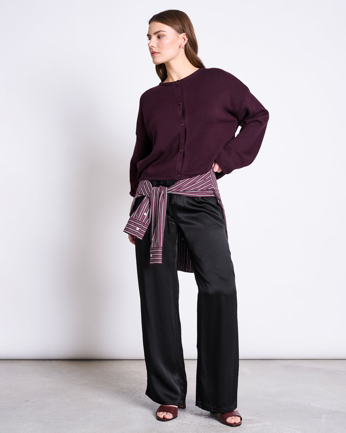 A woman stands against a plain background wearing jan-n-june's LIGHT CARDIGAN ASAS CHERRY LACQUER GOTS, black wide-leg pants, and a striped shirt tied around her waist. She looks to the side with one hand on her hip.