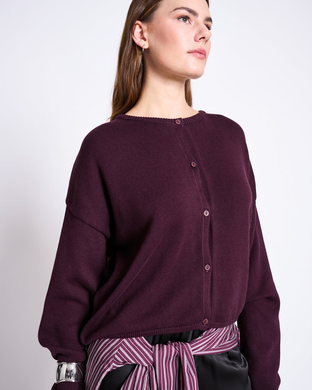 A woman in the jan-n-june LIGHT CARDIGAN ASAS CHERRY LACQUER GOTS, made from GOTS certified organic cotton, with a striped shirt tied at her waist, stands against a plain light background looking slightly to the side.